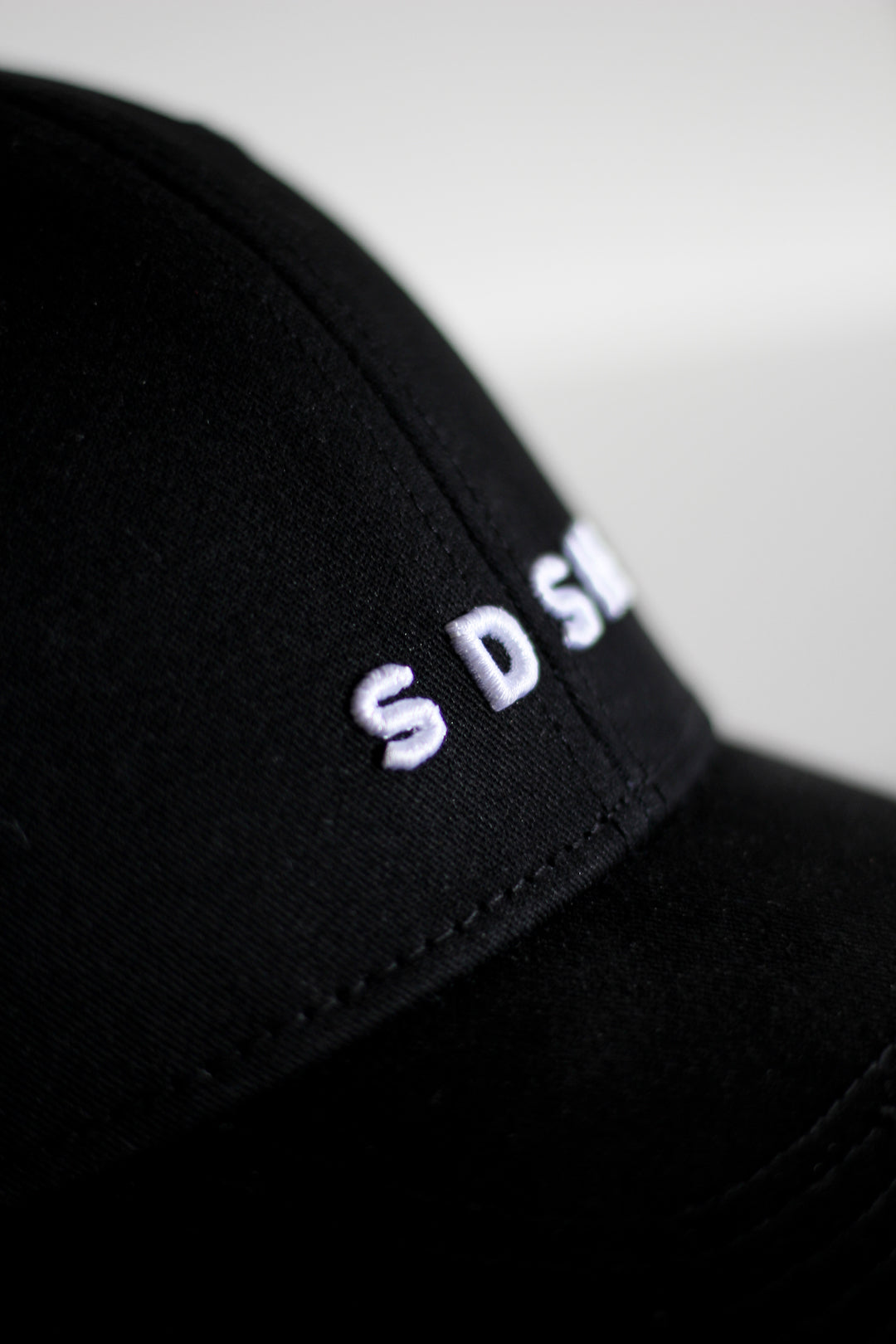 Black cotton baseball cap, 6 panels White 3-D embroidered SDSH monogram curved peak with 6 brim stitches reinforced 2 front panels adjustable fabric strap closure with metal buckle & loop inside printed tape 'FOLLOW YOUR WILD LOVE' 100% cotton twill